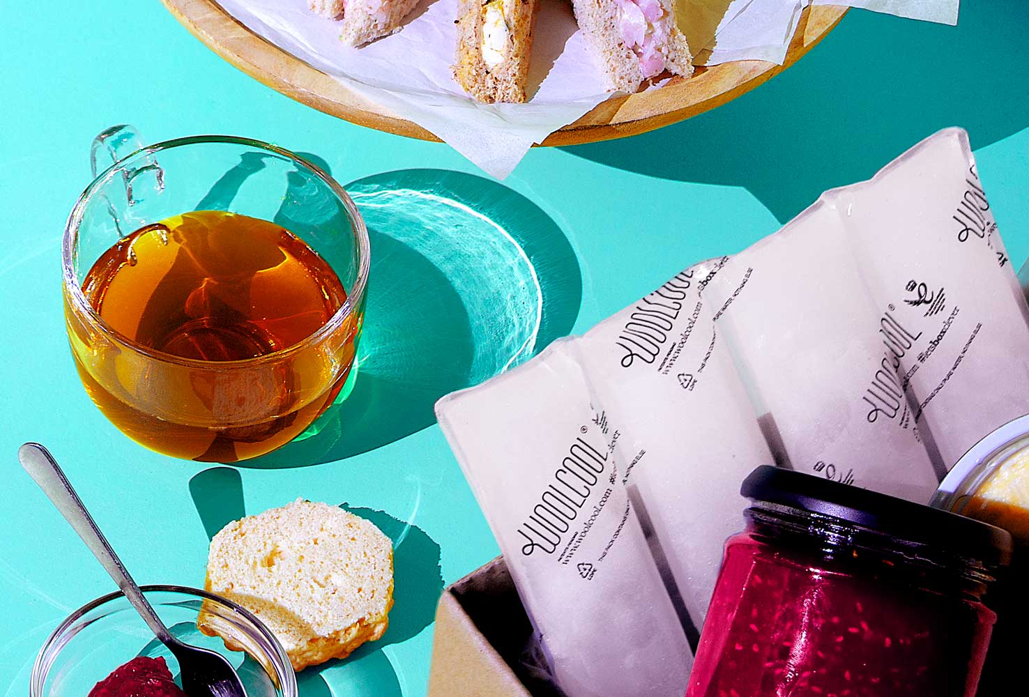 A placement of afternoon tea scone items surrounding a cardboard box. inside the box is a condiment jar (jam) posed on top of a Woolcool branded frozen ice pack.