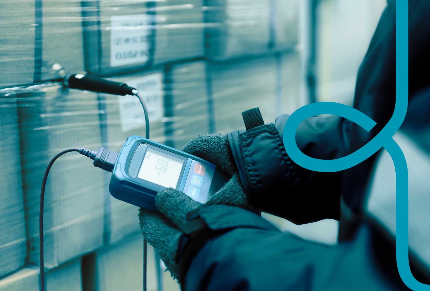 A warehouse operatives hands in frame, wearing protective gloves and holding a temperature monitoring device. In the background are wrapped stacked goods.