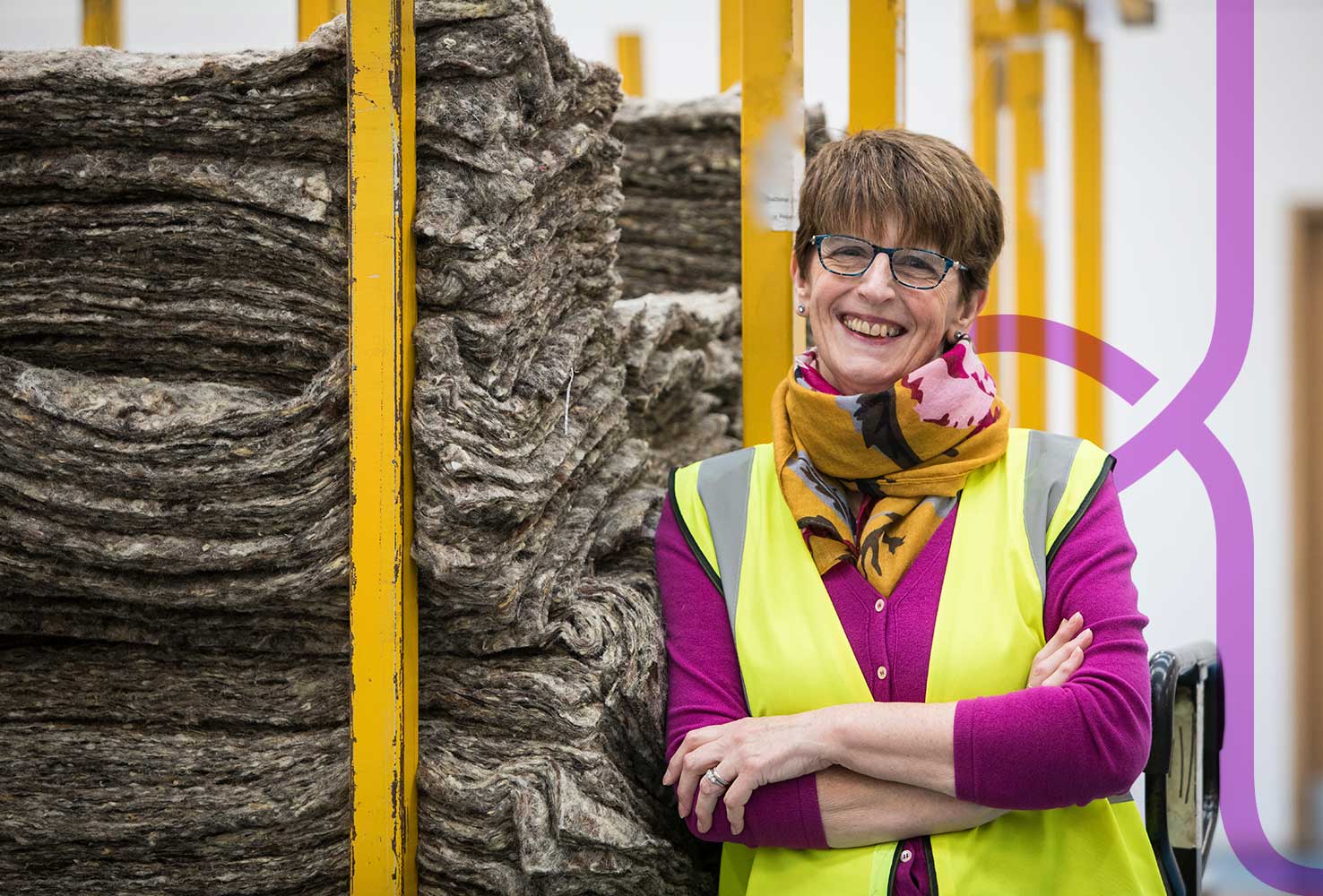 Woolcool founder Angela Morris, standing with her arms folded in a relaxed happy posture and smiling at the camera. She is leaning against a pallet of stack wool fleece